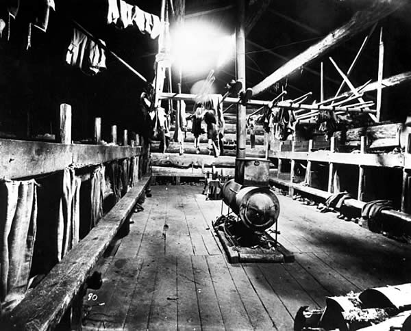 Photo of a bunkhouse interior at a lumber camp: a stove sits in the center, a rack hangs above it with clothes; bunk beds and benches (deacon seats) line the two walls, ca. 1910.