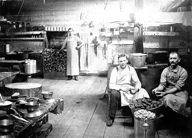 Photo of a lumber-camp kitchen, a male cook stands next to a pile of wood and two male cookees sit peeling potatoes, 1900.