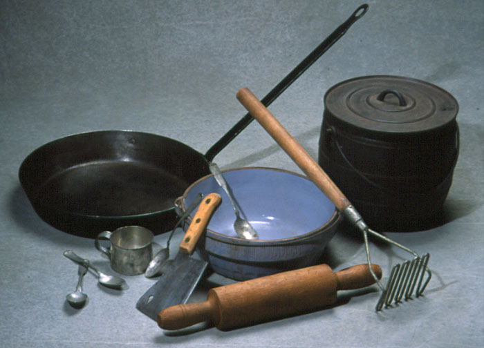 Color photo of lumber camp cooking tools: frying pan, bean pot, large bowl, potato masher, rolling pin, meat cleaver, measuring cup, and spoons.