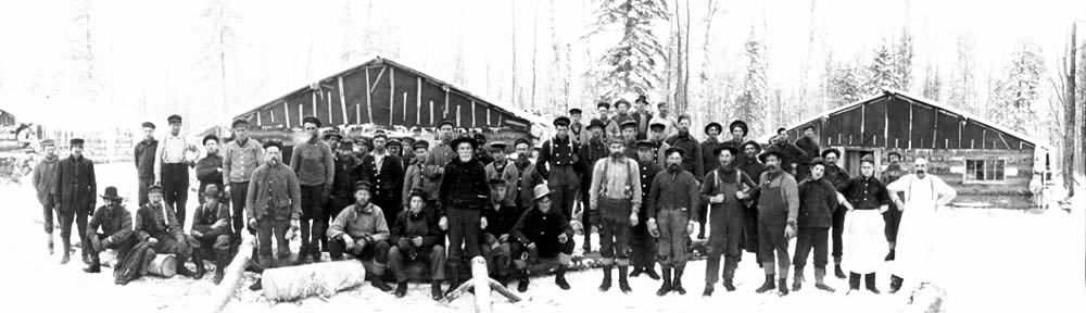 Photo of lumber crew and cooks posing infront of camp buildings, ca. 1900-1920.