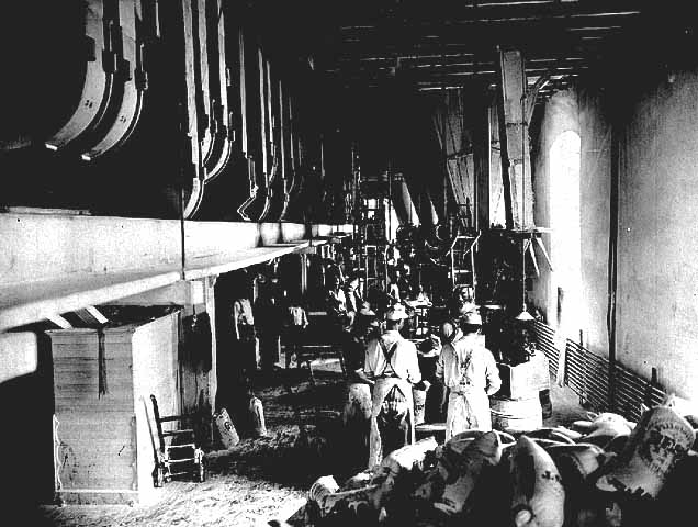 Photo of the flour-packing floor at the Pillsbury A Mill, two workers in foreground wear aprons and hats, 1902.