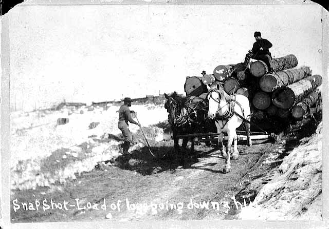 Photo of a load of logs on a sled pulled by two horses, a teamster sits on the load and a man throws straw onto the road to slow the sled, 1885.