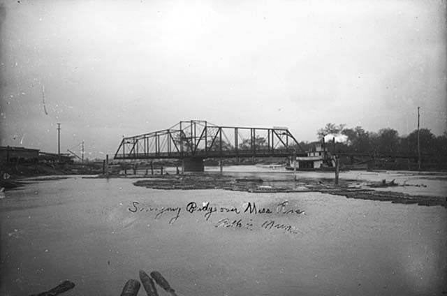Photo of metal swing bridge crossing the Mississippi River in Aitkin showing a steamboat and logs on the river, ca. 1900.