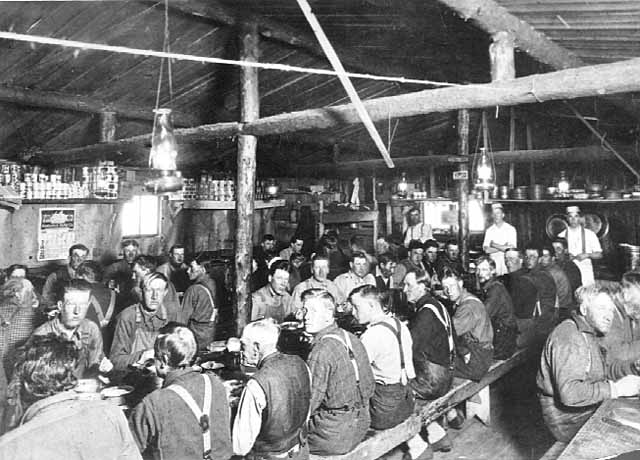 Photo of lumberjacks seated at three long tables in a log-structured cook shack with cooks standing in the background, 1900