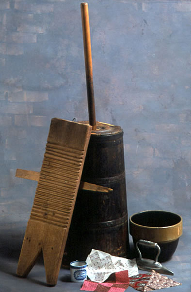 Color photo of a butter churn, washboard, mixing bowl, child's cup, flat iron, and quilt squares.