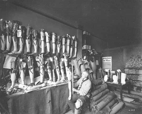 Photo of the Artificial Limb Company's interior: artificial legs hang from the wall and a man wearing a prosthetic sits in the center adjusting a leg, 1918.